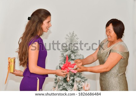 view of a woman hiding a golden wrapped gift box gives a silver wrapped gift box to an asian woman near a Christmas tree. Christmas holiday and gifts concept.