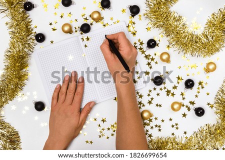 Female hands with pen and notebook among golden and black Christmas baubles, garland and confetti stars. Wish list, New Years resolutions or 2021 plan, planning concept. Party preparation, flat lay