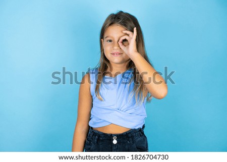 Young beautiful child girl over isolated blue background doing ok gesture shocked with smiling face, eye looking through fingers