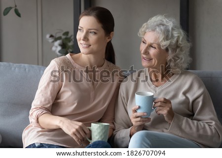 Smiling dreamy mature mother with grownup daughter drinking tea or coffee, holding cups, sitting on couch in living room, elderly woman with granddaughter looking to aside, dreaming about good future Royalty-Free Stock Photo #1826707094