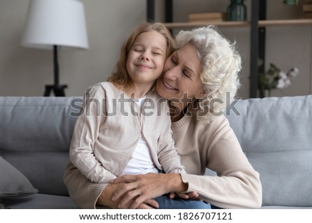 Pretty little girl sitting on happy mature grandmother laps, hugging, enjoying tender moment, good family relationship, smiling elderly woman embracing preschool granddaughter, sitting on cozy couch Royalty-Free Stock Photo #1826707121