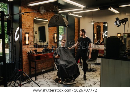 Photosession in a barber shop. Client using phone in a chair, hairdresser next to him looking in the window. All kinds of photo studio equipment around them. Royalty-Free Stock Photo #1826707772
