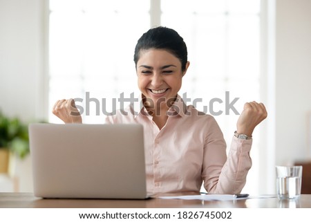 Overjoyed young indian female employee sit at desk in office look at laptop screen read good news online. Happy millennial ethnic woman worker feel excited euphoric with message or notice on computer. Royalty-Free Stock Photo #1826745002