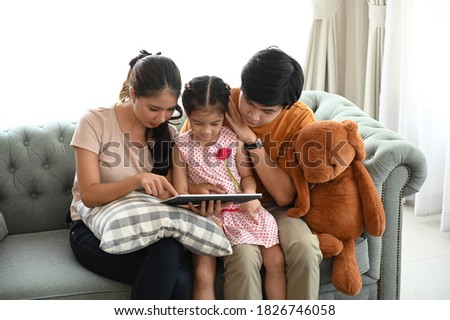 Young joyful casual family sitting on couch and watching funny video or cartoons in tablet.