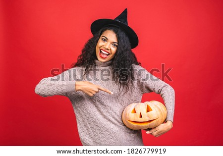 Portrait of Halloween african american witch woman holding a pumpkin and big hat isolated over red background. Halloween style, Halloween party.