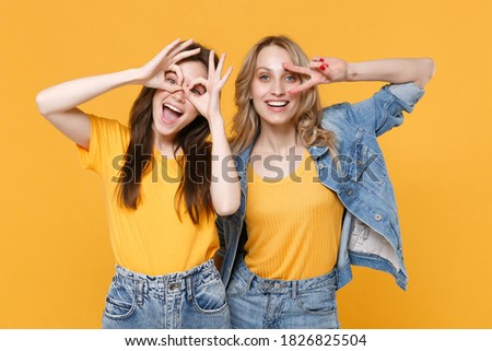 Two funny young women friends in casual t-shirts denim clothes showing victory sign hold hands near eyes imitating glasses or binoculars isolated on yellow background studio. People lifestyle concept