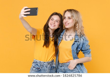 Two smiling pretty young women friends in casual t-shirts denim clothes isolated on yellow background studio portrait. People lifestyle concept. Mock up copy space. Doing selfie shot on mobile phone