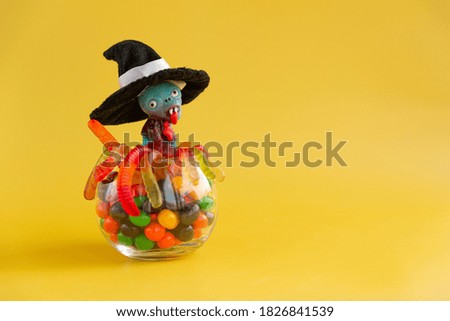 There is a lollipop in the form of a blue zombie in a witch's hat in a round jar with multi-colored candies and jelly worms on a yellow