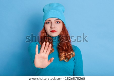 Confident redhead woman keeps palms towards camera, has displeased expression, looks at camera, makes stop gesture, ginger lady gives warning prohibits action, wearing blue sweater and cap.