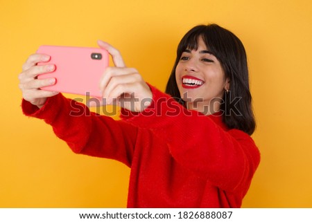 Caucasian brunette woman wearing red casual sweater isolated over yellow background taking a selfie to post it on social media or having a video call with friends.