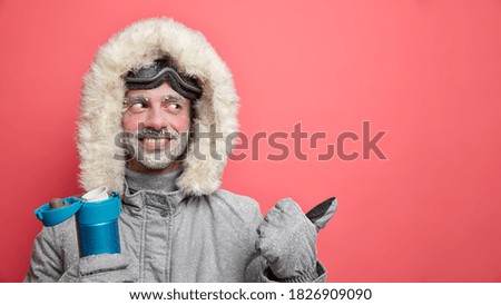 Happy adult man covered by frost wears jacket with fur satisfied after going snowboarding during cold frosty day warms with hot beverage from flask points away on copy space. Winter sports concept