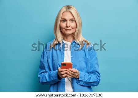 Pleased adorable blonde middle aged woman holds smartphone uses mobile phone application connected to high speed internet dressed in stylish blue shirt stands indoor. People and technology concept