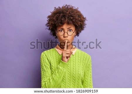 Mysterious serious young Afro American woman makes silence gesture has displeased expression presses index finger to lips dressed in green sweater. Slilence please. Strict female makes hush sign