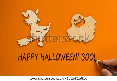 Hand with black pencil writing 'happy halloween, boo'. Wooden witch and ghost. Beautiful orange background. Celebrate concept.
