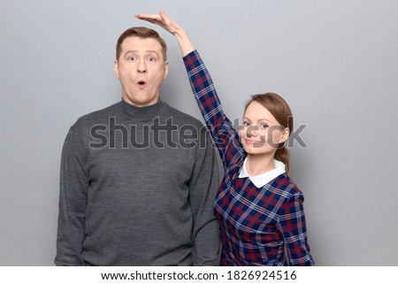 Studio shot of short woman standing and showing height of tall man, woman is smiling, man is amazed, over gray background. Concept of diversity of people's heights, tall and short persons Royalty-Free Stock Photo #1826924516