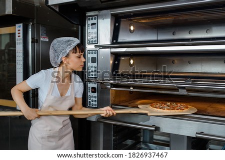 Young caucasian woman baker is holding a wood peel with fresh pizza and put it in an oven at a baking manufacture factory. Tasty pizza  factory concept.
