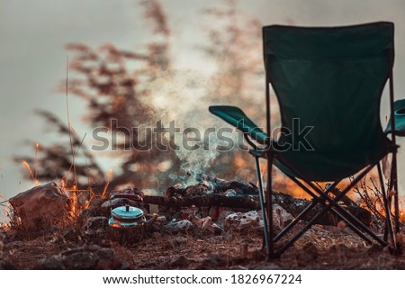 Conceptual Photo of an Autumnal Peaceful Recreation. Empty Camp Chair near Teapot in the Campfire. No People. Tranquility and Relaxation in the Nature.