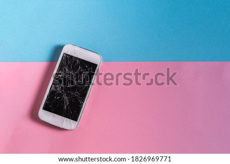Broken mobile phone with cracked display on Blue and pink paper background, outdoor, natural light.