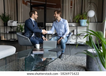 Happy two male partners shaking hands, making agreement after negotiations in modern office. Smiling young leader ceo in suit praising young employee, thanking for good job, achievement concept. Royalty-Free Stock Photo #1826971622