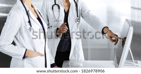 Two female physicians are discussing medical therapy, while standing at the table in a clinic office. Doctors use pc computer at work.  