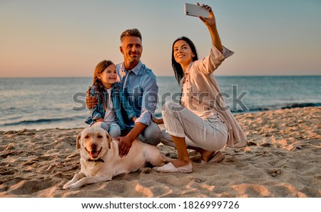 Happy family concept. Young attractive mother, handsome father and their little cute daughter sitting together on the beach with dog and making selfie on a smart phone.