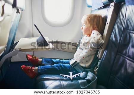 Adorable little toddler girl traveling by plane. Small child sitting by aircraft window and using a digital tablet during the flight. Traveling abroad with kids. Unaccompanied minor concept