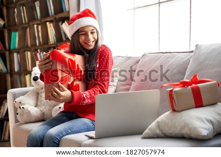 Happy indian child kid girl wearing santa hat holding Christmas gift box, showing New Year present to webcam talking with family, social distance friend by video conference call meeting chat at home.