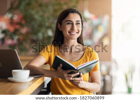 Excited young woman sitting in cafe, using laptop and drinking coffee, holding notepad, copy space. Happy pretty lady with notebook dreaming about successful career, looking for job online