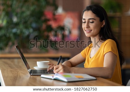 Young business woman planning her day, writing in notepad, sitting in front of laptop, drinking coffee or tea, cafe interior, side view, empty space. Happy arab lady taking notes while websurfing