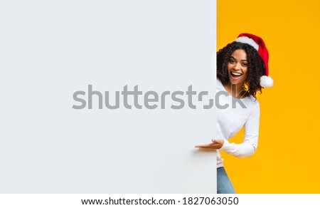 Christmas Offer. Happy African American woman In Santa Hat Pointing At White Advertisement Board With Hand Over Yellow Background, Free Space