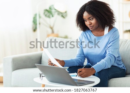 Writing Business Report. Portrait of professional african american woman working at home, holding and reading paper or financial document, sitting on the couch at table with laptop computer