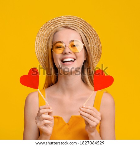 Summer Love. Joyful young girl holding two red paper hearts on sticks in hands, having fun over yellow background, copy space
