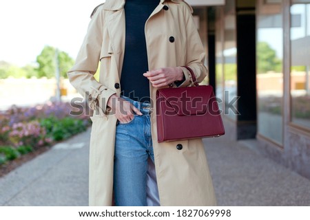 Fashionable young woman wearing beige trench coat, sweater and blue jeans. She is holding burgundy colour leather handbag in hand. Street style.