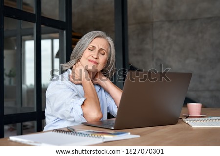 Tired stressed old mature business woman suffering from fibromyalgia neckpain working in office sitting at table. Overworked senior middle aged lady massaging neck feeling hurt pain from sedentary job Royalty-Free Stock Photo #1827070301