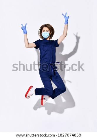 Young positive woman doctor dentist in dark blue uniform, gloves and protective medical mask jumping and showing peace sign with fingers over white wall background. Professional woman dentist concept