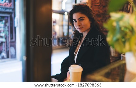 Half length portrait of attractive female customer looking at camera during coffee break in sidewalk cafe, beautiful teenage Caucasian girl 20 years old posing during resting time in street cafeteria