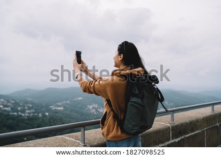 Back view of female traveler with backpack making photo on mobile phone during trip on vacations, rear view of woman tourist taking picture of nature environment exploring destinations in journey