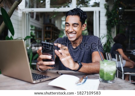 Cheerful adult Hispanic guy in gray shirt with watch focusing on screen and using smartphone while sitting at laptop with notepad glasses and green drink in cafe during daytime on blurred background