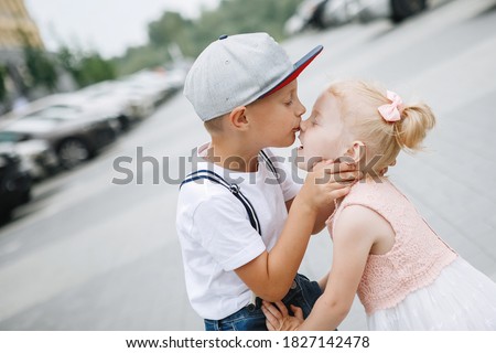 Boy and a girl are kissing on a city street. A blonde with ponytails in a pink dress and a child in a baseball cap.