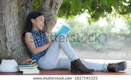 little asian girl reading a book under big tree with hat, book, and model plane beside. children and science. blurred background. learning the imagination and dreams of rural child. studying at home.