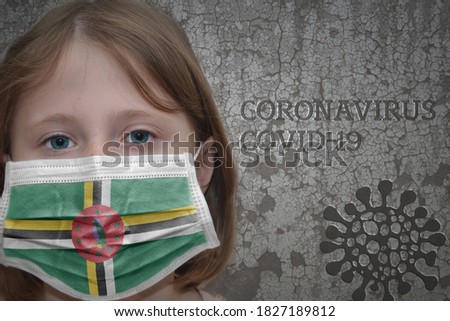 Little girl in medical mask with flag of dominica stands near the old vintage wall with text coronavirus, covid, and virus picture. Stop virus