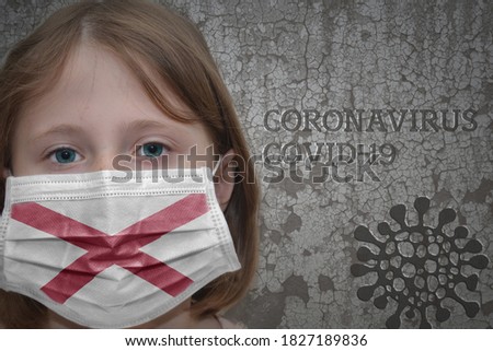 Little girl in medical mask with alabama state flag stands near the old vintage wall with text coronavirus, covid, and virus picture. Stop virus