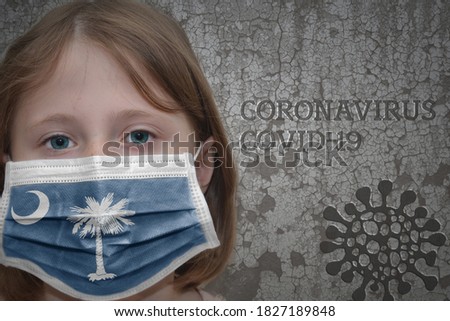 Little girl in medical mask with south carolina state flag stands near the old vintage wall with text coronavirus, covid, and virus picture. Stop virus