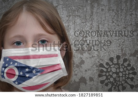 Little girl in medical mask with ohio state flag stands near the old vintage wall with text coronavirus, covid, and virus picture. Stop virus
