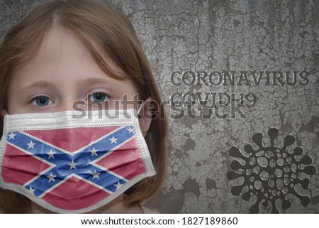 Little girl in medical mask with confederate jack flag stands near the old vintage wall with text coronavirus, covid, and virus picture. Stop virus