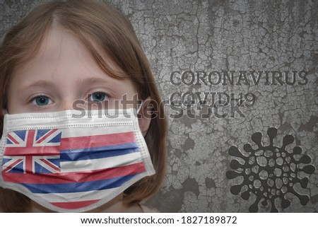 Little girl in medical mask with hawaii state flag stands near the old vintage wall with text coronavirus, covid, and virus picture. Stop virus