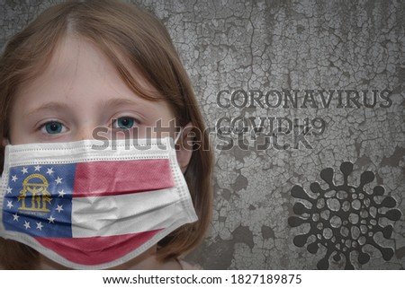 Little girl in medical mask with georgia state flag stands near the old vintage wall with text coronavirus, covid, and virus picture. Stop virus