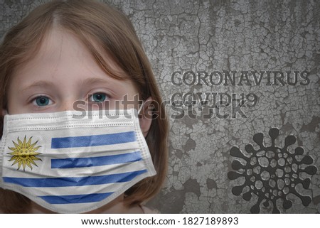 Little girl in medical mask with flag of uruguay stands near the old vintage wall with text coronavirus, covid, and virus picture. Stop virus