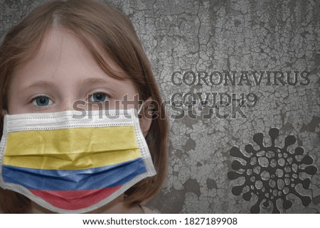 Little girl in medical mask with flag of colombia stands near the old vintage wall with text coronavirus, covid, and virus picture. Stop virus