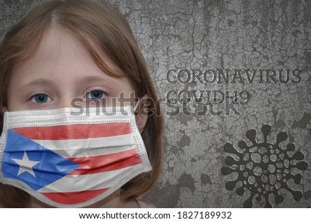 Little girl in medical mask with flag of puerto rico stands near the old vintage wall with text coronavirus, covid, and virus picture. Stop virus